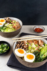 Two bowls of ramen soup with chicken breast, vegetables, mushrooms, pak choi and egg on a gray napkin and on a marble table with chopsticks. Top view, healthy eating concept.