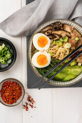 Bowl of ramen soup with chicken breast, vegetables, mushrooms, egg, chopsticks and a gray napkin on a slate stone on a white wooden table . Top view, healthy eating concept