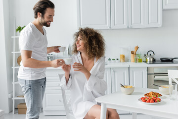 happy young man giving cup of coffee to curly girlfriend during breakfast in kitchen.