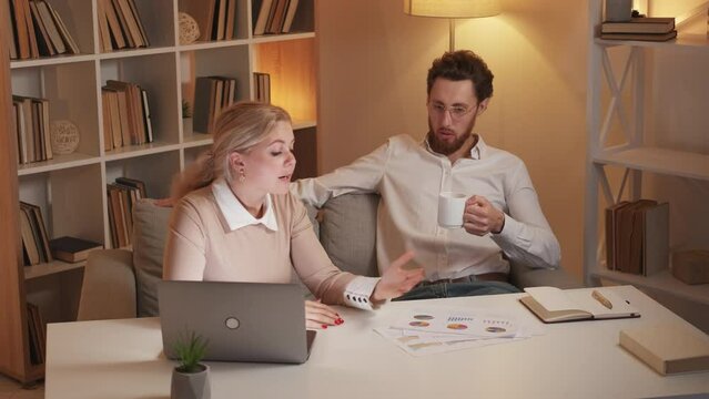 Bad work. Displeased woman. Colleagues quarrel. Angry female manager showing wrong documents to relaxed careless man sitting sofa drinking coffee in light room interior.
