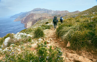 zwei wanderer bei starker wind an der Küste Mallorcas auf dem Weg der Tramuntana mit Kalk Klippen in der Ferne und traumhafte Landschaft mit dem Mittelmeer und satt grüne Flora im März 2022