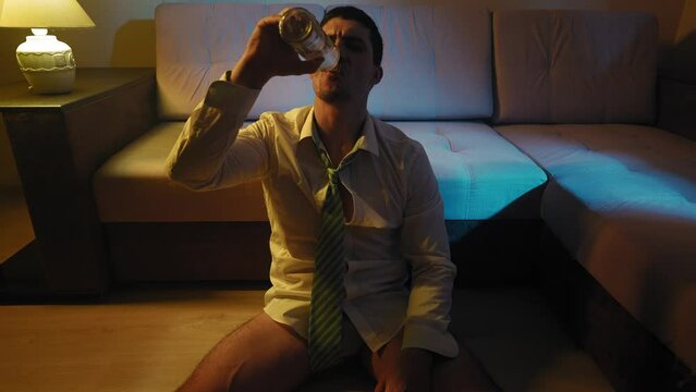 Upset Alcoholic Man Without Trousers in a White Shirt and Tie Sits on the Floor of the House and Gets Drunk with Alcohol from a Glass Bottle at Night. Concept of Alcoholism and Depression. Slow Motion