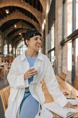 View of dark-haired attractive woman looking away in hands cup coffee. Staying in cafe, drinking after work, wearing casual clothes. Leisure of life concept