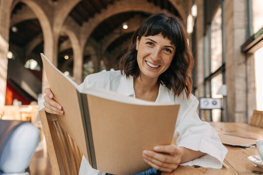 Picture of pretty brunette cute woman luffing and look at camera indoors. Wearing white shirt, holding notebooks sitting on the chair. Positive emotion concept 