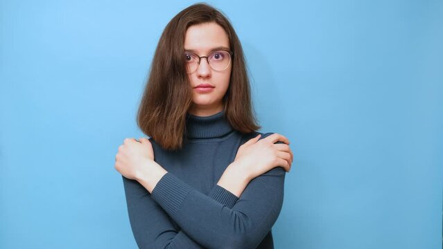 Displeased brunette student girl in glasses and a sweater embraces herself, holds shoulders, feels cold, missed opportunity, expresses regret, dreams of warm and cozy home on blue background