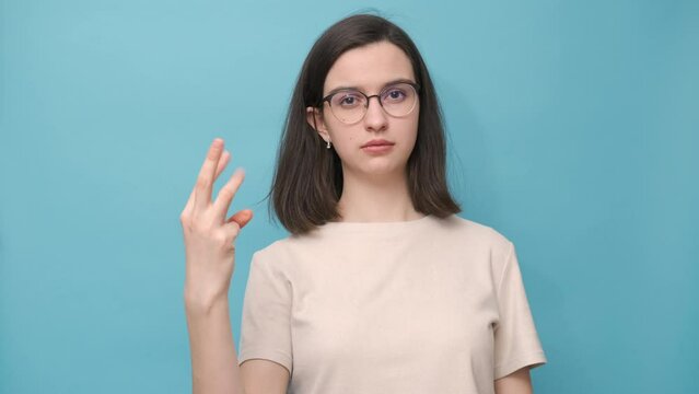 Portrait of a student girl with glasses and a beige blouse unbending her fingers in turn, counting on her fingers on a blue background, 4K. Nonverbal communication. Mathematics