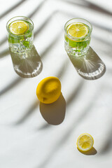 Two glasses of cocktail or water with sliced lemon and mint on white sunlit background with palm...