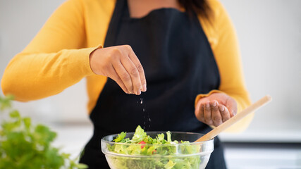 Millennial african american woman in apron salting salad, cooking dinner at table with organic fresh vegetables