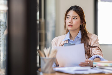 Image of young beautiful brooding Asian woman working with laptop while sitting at laptop in office, thinking of professional plan, project management, considering new business ideas.