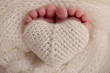 The tiny foot of a newborn baby. Soft feet of a new born in a wool white blanket. Closeup of toes, heels and feet of a newborn. Knitted heart in the legs of baby. Macro studio photography. 