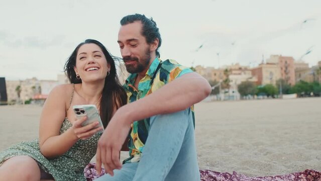 Romantic couple uses mobile phone while sitting on the beach on buildings background