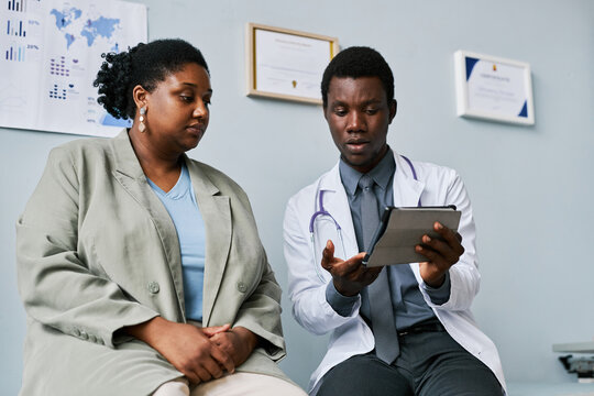 Low Angle Young Black Doctor Patient Talking In Clinic Looking At Tablet Screen