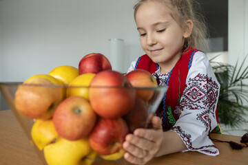 Fototapeta na wymiar A little girl in a national Ukrainian costume stands at the table and holds a vase with bright apples in her hands.