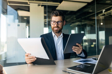 Cheerful middle aged businessman with documents and tablet working in office interior, free space