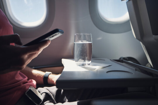Glass ofwater on table of seat in airplane. Passenger is using phone during flight..