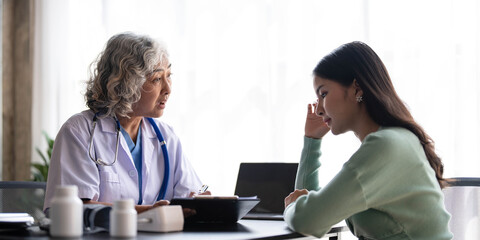 Woman senior doctor is Reading Medical History of Female Patient and Speaking with Her During Consultation in a Health Clinic. Physician in Lab Coat Sitting Behind a Laptop in Hospital Office.