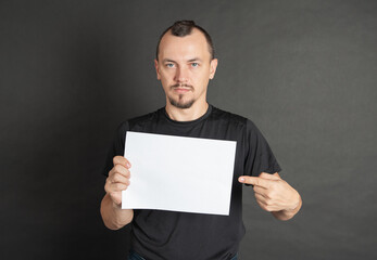 Young handsome man holding empty white sheet of paper and showing on it by his finger