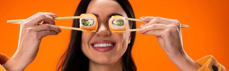 cheerful brunette woman covering eyes with fresh sushi rolls isolated on orange, banner.