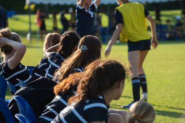 Substitute (bench, backups, interchange, reserves) girl players watching their team playing on a sideline waiting for their turn to play in an outdoor sport: soccer/football, rugby tournament.
