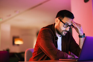 Tired man working over the laptop, sitting at the workplace, with glasses on.