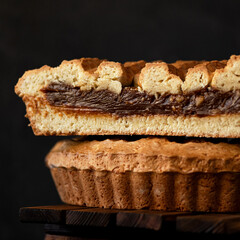 American pie with chocolate filling and mesh top. Homemade holiday pastries for family tea drinking. Sand cake. Dessert in cut form on wooden table. Dark background. Close-up. Side view