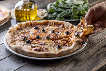 A man's hand holds the first piece of freshly baked pizza, fresh basil and a carafe of olive oil in...