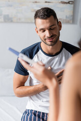 Smiling man in pajama standing near girlfriend with pregnancy test in bedroom.