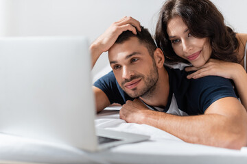Smiling woman hugging boyfriend near blurred laptop on bed.