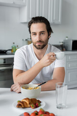 Fototapeta na wymiar bearded man holding cup of coffee near tasty breakfast on table in kitchen.
