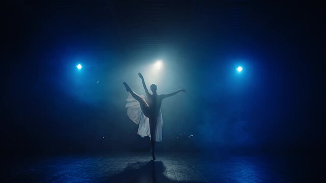 Silhouette of graceful ballerina in blue backlight of spotlights. Young ballet dancer twirling on toes in pointe shoes in stage smoke