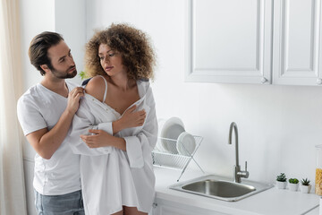 bearded man undressing curly woman in white shirt in kitchen.