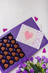 Violet box of heart shaped chocolates, postcard and flowers on white background. Valentines day 14 February sweet present
