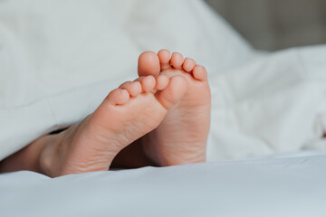 View on kid bare feet on white bed. Bedroom lit with morning light. Child sleeps in pajamas comfort
