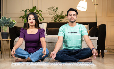Peaceful Young indian couple doing meditation or yoga with eyes closed while at home during early...