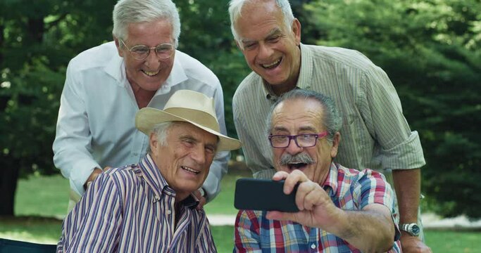 A Group of Senior Men Enjoying the Weather Outdoors in a Park. Elderly Male Friends Jokingly Making Silly Faces while Taking Photos on a Smartphone. Active and Lively Senior Citizens Using Technology
