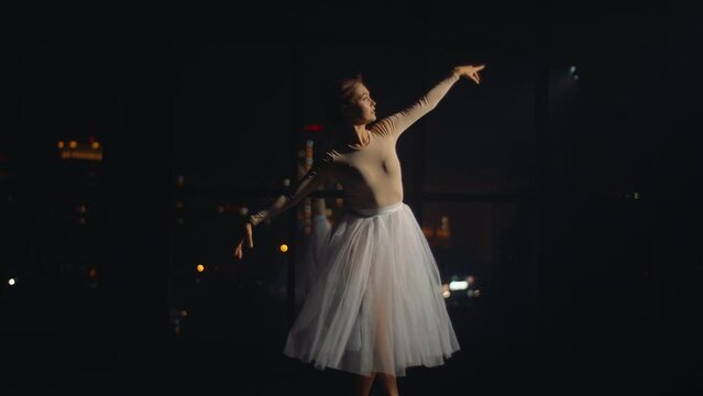 Young graceful ballerina dances on stage against backdrop of lights of night city. Beautiful ballet dancer spins and dances