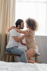 side view of bearded man sitting on bed bench and hugging sexy girlfriend in bedroom.