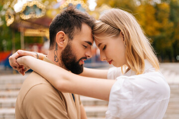 Naklejka premium Close up of romantic young couple in love touching each other foreheads hugging standing with closed eyes in park in summer day. Handsome bearded man and blonde woman enjoying weekend outdoors.