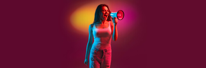 Emotive young girl in casual clothes shouting in megaphone over gradient dark pink background in neon light. Information, news. Concept of emotions, facial expression, youth. Banner, flyer, ad