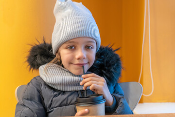 Young girl in hat and warm clothes enjoying hot cacao drink with straw.