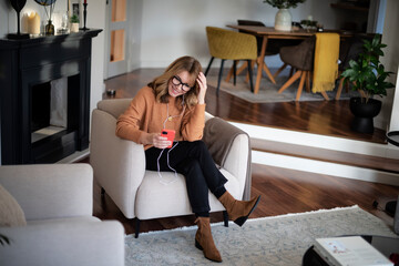 Obraz premium Happy woman using earphones and cellphone while sitting in an armchair at home