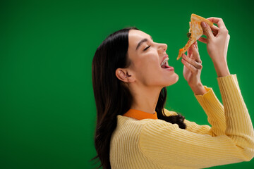 side view of young and hungry woman eating piece of pizza isolated on green.