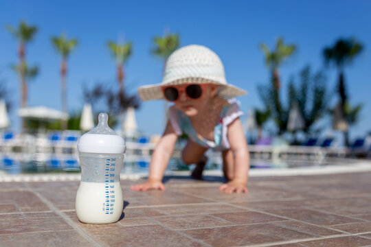 Bottle with milk and fashionable baby girl crawling in the background near swimming pool. Children and summer vacation concept.