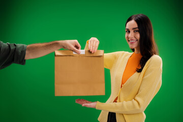 pretty brunette woman taking paper bag from delivery man and smiling at camera isolated on green.