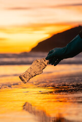Hand with a glove picks up a plastic bottle from the shore. Vertical photo. Orange reflection of the sunset on the water. Concept Climate change, ecology, volunteering.