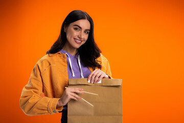 Smiling brunette woman holding bamboo chopsticks and paper bag isolated on orange.