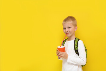 blond boy with a school backpack drinks through a straw from a glass on a yellow background with copy space, back to school, school meals