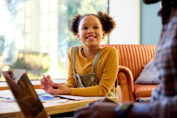 Father At Home With Laptop With Daughter In Lounge As She Does Homework At Table