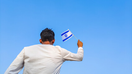 Boy holding Israel flag against clear blue sky. Man hand waving Israeli flag view from back, copy space