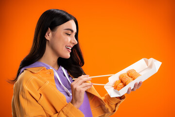 Smiling brunette woman holding takeaway sushi and chopsticks isolated on orange.